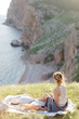 © NesolenayaAleksandra - Picnic on the grass on mountain over sea view on sunny summer day. Young beautiful woman, wrapped in a blanket, sitting on the plaid with fruits, drinking tea and relaxing on nature. Back view.