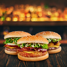 Three Burgers At Brown Wooden Tabletop With Blurred Bar At Backdrop . Fastfood Concept With Hamburgers At Wooden Table