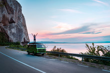 Young Girl Standing On Top Of Vintage Classic Camper Van With Hands Wide Open. Freedom On The Summer Road Trip Beside Seashore With Beautiful Sunset. - Image