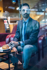 Wall Mural - Handsome young south asian indian business man in formalwear having a meal in cafe during coffee break. Through window view.