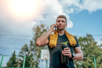 Wall Mural - A male athlete, summer city after a workout, calls on phone, holds in his hand a shaker, a bottle of water, a towel around his neck. Resting after a hard workout. Active lifestyle, workout in nature.