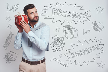 Wall Mural - What is inside? Attractive stylish man is holding a gift box near his ear and trying to guess what is inside while standing against grey background with different doodle illustrations on it