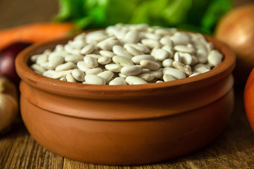 Wall Mural - Uncooked white bean in a clay bowl on wooden table. Tomato, red onion, garlic, carrot and lettuce in the background.