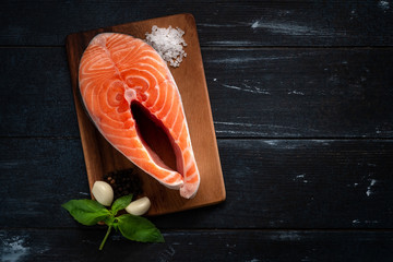 Wall Mural - Raw salmon steak on a wooden Board with salt and basil on a black background. Fresh red fish. Healthy and diet food. Top view.