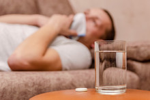 Man Sitting On The Couch. He Has A Runny Nose, So He Blows Her Nose And Wipes Away Tissue Paper. Place The Medicine And A Glass Of Water On The Table Next To It. The Concept Of The Disease