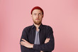 Photo of questioning bearded man in basic shirt, with red hat, with folded arms, look up with one raised eyebrow over pink background.