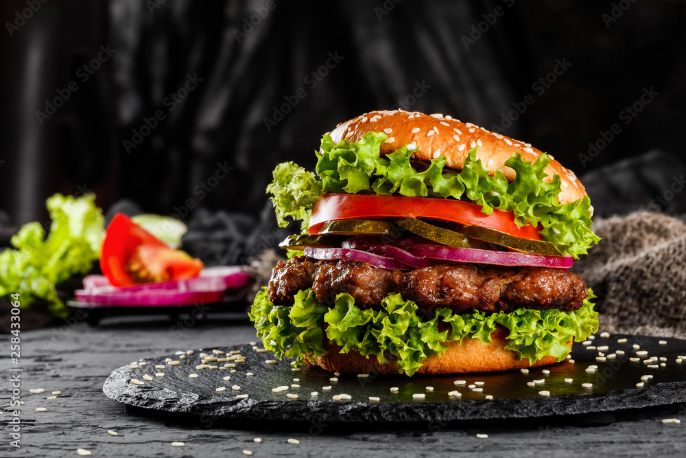 Beef burger with tomatoes, red onions, cucumber and lettuce on black slate over dark background. Unhealthy food - obrazy, fototapety, plakaty 
