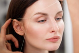 Fototapeta Zwierzęta - Cosmetologist plucks client eyebrows by thread. Close up