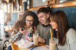 © dusanpetkovic1 - Three smiling multicultural classmates sitting at coffee shop, drinking coffee and looking at smart phone. On desk notebooks, markers and pens.