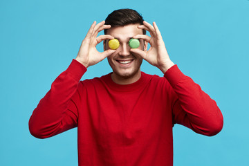 Food, pastry, joy and fun concept. Picture of funny unshaven young brunette male in red sweatshirt posing isolated at blue studio wall, holding two round almond cookies at his eyes and smiling happily