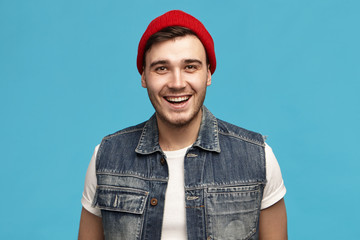 Wall Mural - Positiveness, joy and happiness concept. Picture of unshaven friendly joyful young European man in red headwear and jeans vest smiling broadly being in good mood enjoying carefree time of his youth