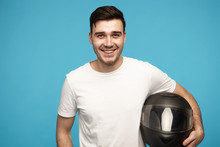 People, Active Lifestyle, Sports And Extreme Concept. Waist Up Image Of Happy Friendly Looking Guy With Black Hair Posing In Studio With Black Safety Motorcycle Helmet Under His Arm, Smiling Broadly