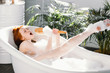 Vivacious emotional young woman with red hair bun taking bath at home, being in good temper, singing a song holding body brush as a microphone, giving way to her talant and joy