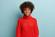 Negative Emotions And Facial Expressions Concept. Black Young Woman Frowns Face And Clenches Teeth, Sees Something Digusting, Annoyed By Bad Suggestion, Wears Red Winter Sweater, Shows Displeasure