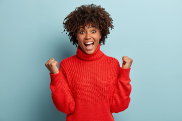 Poster - Glad young woman shows gesture of victory, wins competition, keeps fists raised, achieves desired result, screams from joy, wears red jumper, models over blue background. Reaching goal or aim