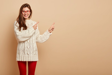 Horizontal shot of positive European woman with cheerful expression, points away, shows blank space for your advertisement or promotion, dressed in casual white jumper, advertises something.