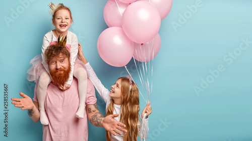 International childrens day concept. Fatigue bearded male entertainer at kids party, amuses girls, gives piggyback, looks clueless as doest know which game to suggest. Small child with balloons