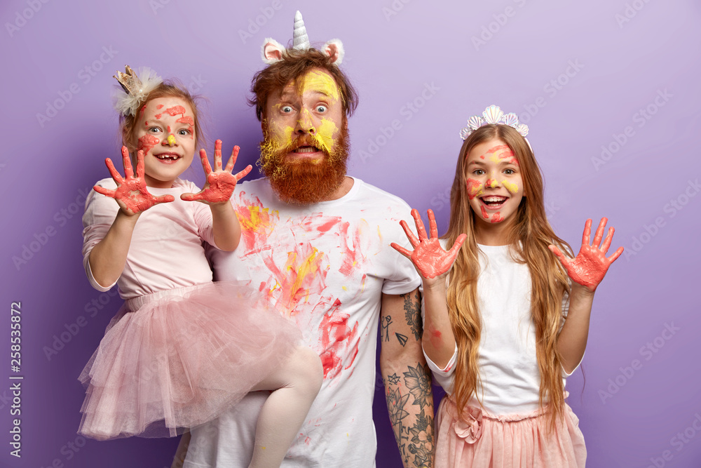 Funny stupefied dad and two adorable daughters show dirty palms with paints, stare at camera together, wear festive carnival costumes, celebrate Children Day, isolated over purple background - obrazy, fototapety, plakaty 