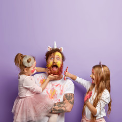 Shocked dad spends free time with daughters, dressed in unicorn costume, two children touch his face and dirty with colourful paints, surprised to be in colors. Father and little girls have fun indoor