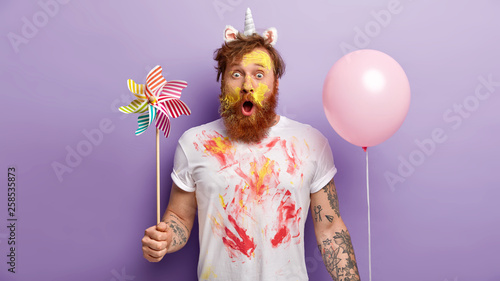 Fotografía Indoor shot of surprised red man stares with shock, holds balloon and windmill i