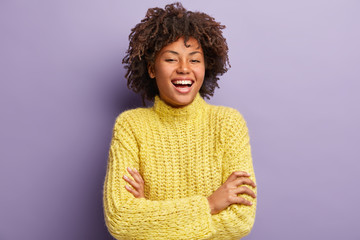 Sticker - Young dark haired young Afro American woman laughs at good joke, keeps arms folded, wears knitted yellow jumper, expresses sincere emotions, has kind expression, isolated over purple background.