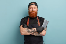 Portrait Of Handsome Professional Male Butcher Has Arms Crossed, Holds Metal Sharp Cleaver, Wear Black Uniform, Ready To Start Work, Isolated Over Blue Background. People And Occupation Concept