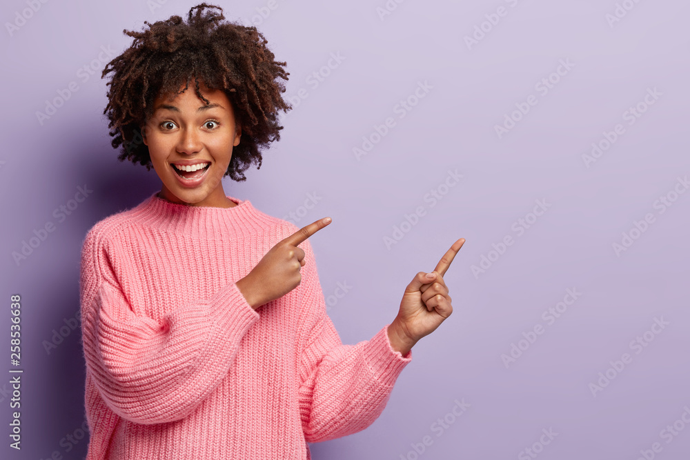 Waist up shot of positive lovely woman with Afro hairstyle, points away with both fore fingers, shows somethig on blank space, has joy expression, wears loose pink jumper, gestures over violet wall - obrazy, fototapety, plakaty 