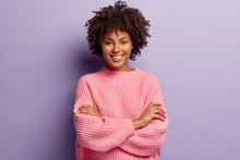 Photo Of Self Confident Pleased Dark Skinned Woman Keeps Arms Folded, Smiles Gladfully While Watches Funny Programme, Has Natural Appealing Beauty, Wears Oversized Pink Jumper, Poses Indoor.