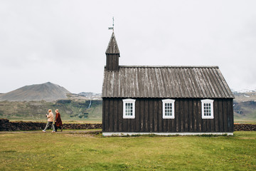 A couple is near a black wooden church in Iceland. Budir Church.