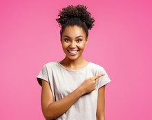 Overjoyed Girl Points Aside, Shows Something At Freespace. Photo Of African American Girl Wears Casual Outfit On Pink Background. Emotions And Pleasant Feelings Concept.