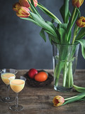 Fototapeta Tulipany - Colorful Easter eggs in nest, two glasses of egg nog and tulips in vase on wooden table.