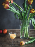Fototapeta Tulipany - Colorful Easter eggs in nest, glass of egg nog and tulips in vase on wooden table.