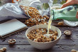 © insta_photos - Female hand holding glass bottle pouring milk in cereal granola flakes bowl with nuts seeds raisins on brown wooden table background, healthy breakfast lifestyle concept, home muesli food oat meal