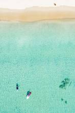 Stunning Aerial View Of Two Unidentified People Doing Stand Up Paddle Board And Windsurf On A Clear And Turquoise Sea While A Woman Sunbathing On A White Beach. Surin Beach, Thailand.