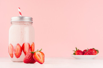 Light dairy strawberry drink in trendy jar with ripe pieces berry on saucer, red striped straw, silver lid on gentle pink background and white wood table.