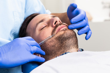 Handsome young man is getting a rejuvenating facial injections. He is sitting calmly at clinic. The expert beautician is filling male wrinkles by hyaluronic acid.