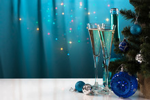 Blue Bottle And Two Champagne Glasses Are Standing On A Light Reflective Surface Near The New Year Tree Decorated With Toys On A Blue Background.