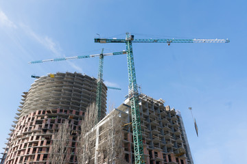 high-rise construction crane with a long arrow of yellow color against the blue sky over a new multi-storey building of concrete and brick under construction