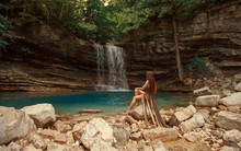 Mysterious River Nymph On Bare Rocks Near Small Blue Lake In Martvili Canyon Lagoon And Wonderful Waterfall, Girl With Red Hair In Long Brown Dress With Long Train Cut. No Model Face, Creative Colors