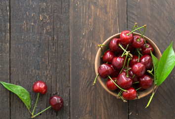 White cup with ripe berries of yellow and red sweet cherries and several fruit with green leaf of cherry tree on wooden background