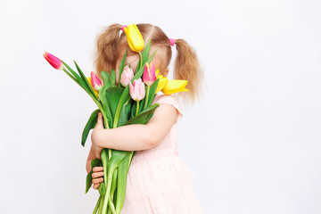 Little beautiful girl blonde with a bouquet of tulips on a light background. Baby girl smiling. Spring and women's day concept. Little girl holding a bouquet of colored tulips. Сopy spase