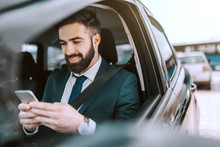 Smiling Caucasian Businessman In Formal Wear Using Smart Phone While Sitting In His Car On Parking Lot.