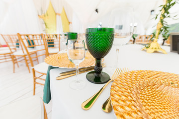 Golden dishes, green wine glasses and napkins. Catering concept. Interior of tent for wedding dinner, ready for guests. Served round banquet table.