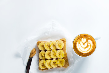 Wall Mural - Healthy vegan breakfast concept. Cappuccino, soy milk foam, peanut butter sandwich, toasted bread, slices of banana. Clean eating. White table background. Top view, close up, copy space, flat lay.