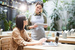 © Seventyfour - Content friendly waitress in apron using tablet for work and asking guest about food and drink preferences, businesswoman working in cafe