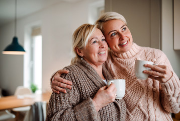 Wall Mural - A senior woman and adult daughter with coffee at home, looking out of window.