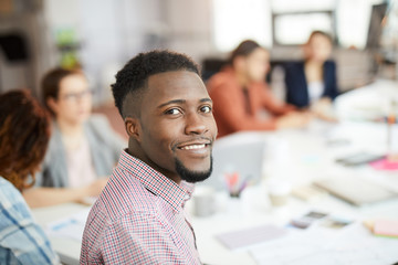 Wall Mural - Head and shoulders portrait of young African-American man smiling at camera while sitting at table in business meeting, copy space