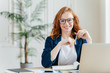 © VK Studio - Portrait of happy redhaired woman employee in optical glasses, has satisfied expression, works with modern gadgets, waits for meeting with colleague, prepares accounting report, sits in own cabinet