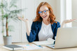 Image of cheerful redhead businesswoman watches webinar or tutorial video, uses free internet connection, raises palms with hesitation, sits at desktop with papers. Female owner of small business