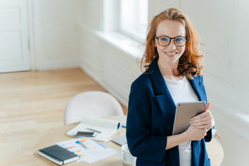 Prosperous female leader of working team holds digital tablet device, develops business ideas, has toothy smile, red hair, wears elegant outfit, stands in own cabinet, involved in working process